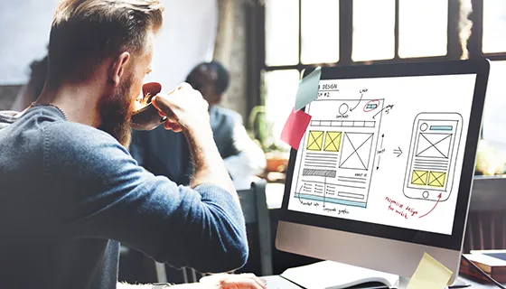 Person drinking coffee while reviewing a website wireframe design on a computer screen, suggesting a web design or development workflow