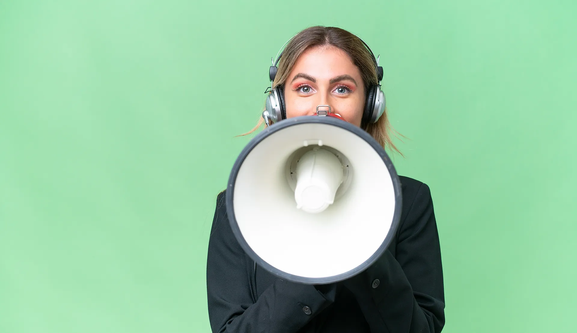 A woman wearing headphones and holding a megaphone in front of her face, with a green background. The focus is on the megaphone, suggesting she is making an announcement or amplifying a message.