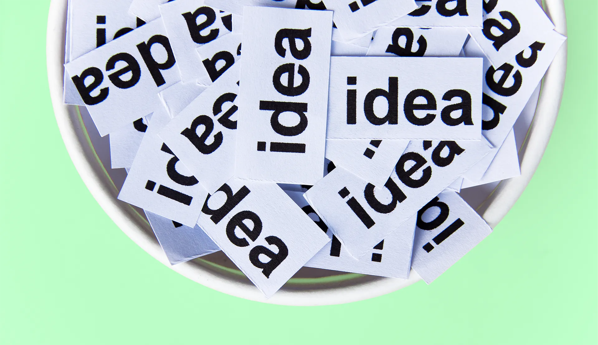 A bowl filled with multiple small paper slips, each displaying the word 'idea' in bold text, against a light green backdrop, symbolizing creativity and brainstorming.