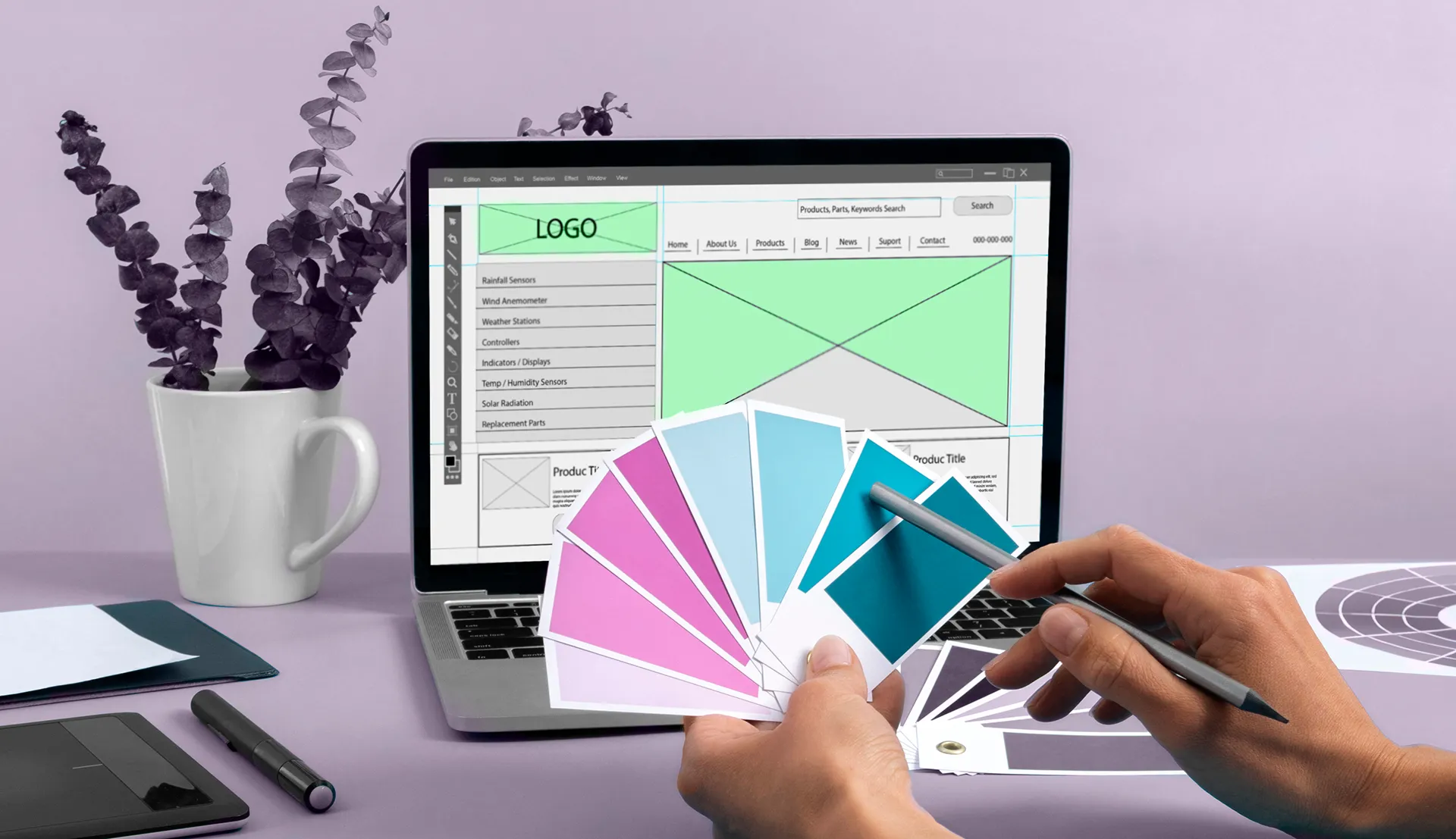 A person holding color swatches in blue, pink, and purple shades while working on a web design wireframe displayed on a laptop. The desk includes a graphics tablet, a pen, papers, and a mug with purple leaves.